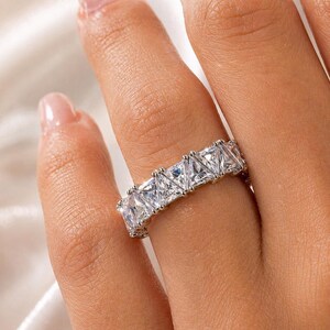 Puede incluir: Un anillo plateado con una fila de piedras preciosas claras, de corte cuadrado. El anillo se lleva en un dedo, resaltando las piedras brillantes. El dise&ntilde;o es sencillo pero elegante, adecuado para el uso diario o para ocasiones especiales.