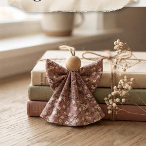 Könnte beinhalten: Ein nähfreier Stoffengel-Anhänger mit einem Kleid mit altrosa Blumenmuster und einem runden Kopf aus hellem Holz. Der Anhänger hat eine Schlaufe aus Jutekordel zum Aufhängen. Der Text lautet No-Sew Fabric Angel Ornament und Easy Beginner PDF Pattern.