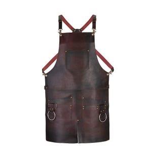 Könnte beinhalten: Eine dunkelbraune Lederschürze mit einer Brusttasche, zwei großen unteren Taschen und zwei an der Vorderseite befestigten Metallringen. Die Schürze verfügt über verstellbare rote Riemen, die sich am Rücken kreuzen und mit Metallclips befestigt sind.