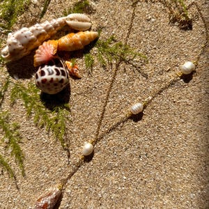 May include: A gold-colored necklace with three white pearls and a teardrop-shaped shell pendant. The necklace is displayed on a sandy surface with several seashells and green sprigs.
