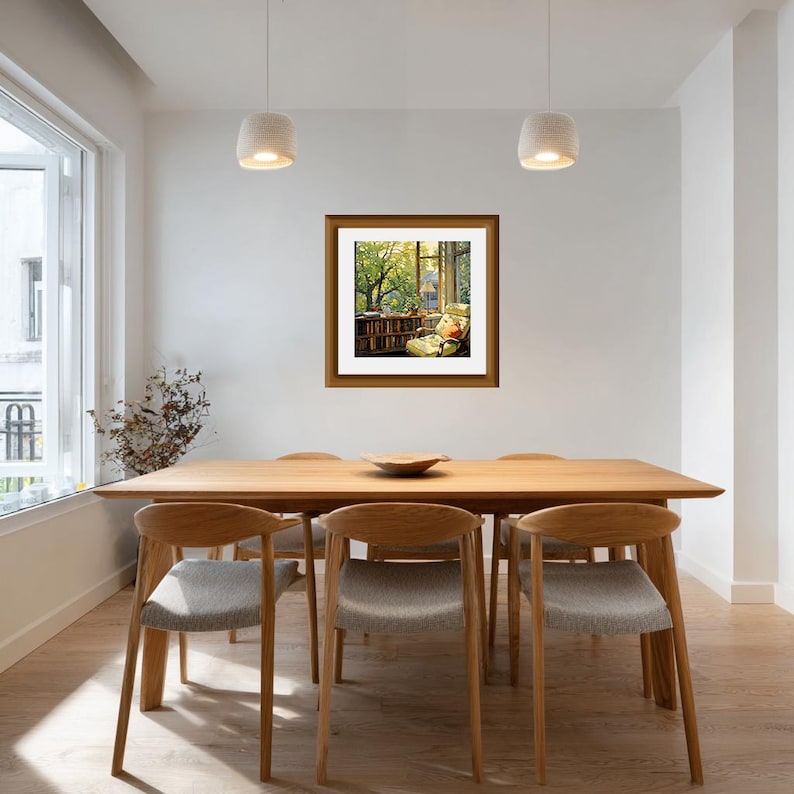 May include: A framed artwork depicting a cozy reading nook with a window view of a lush green tree. The artwork is displayed above a wooden dining table with six chairs. The room has white walls and pendant lights.