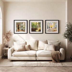 May include: Three framed art prints with black frames hang above a cream-colored sofa. The prints depict interior and exterior scenes with natural light. A small side table with a vase of dried flowers sits to the left of the sofa.