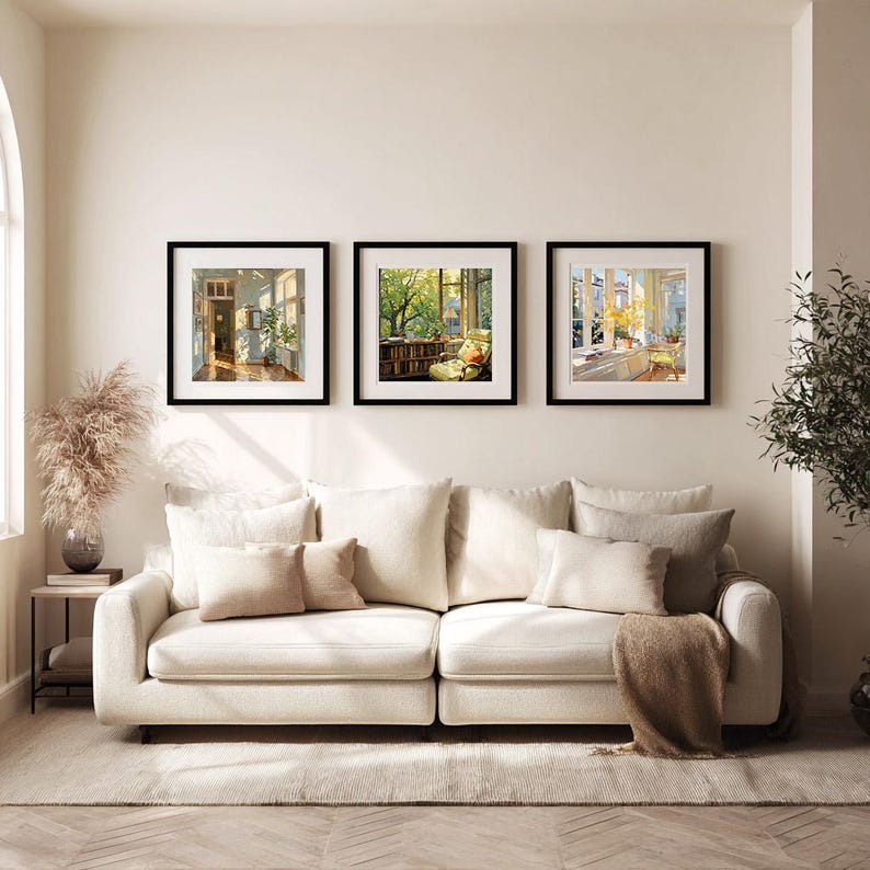 May include: Three framed art prints with black frames hang above a cream-colored sofa. The prints depict interior and exterior scenes with natural light. A small side table with a vase of dried flowers sits to the left of the sofa.
