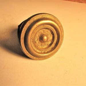 May include: A brass-colored cabinet knob with a circular design. The knob features concentric rings and a central, raised button. The outer ring has a detailed, textured pattern. The knob is likely for furniture or cabinetry.