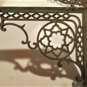 May include: Ornate, dark bronze-colored metal shelf brackets. The brackets feature a decorative openwork design with diamond and floral patterns. The corner bracket has a circular floral design in the center. The brackets are designed to be mounted on a wall.