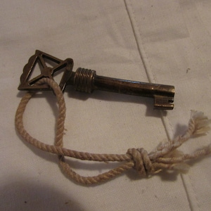 May include: A brass key with a decorative head and a rope attached to it. The key is lying on a white surface.