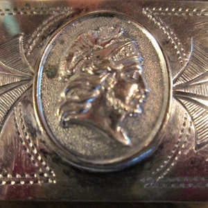 May include: A silver-toned metal belt buckle with an oval central design featuring a profile of a person wearing a headdress. The buckle has an engraved design with a series of lines and curves radiating from the central oval.