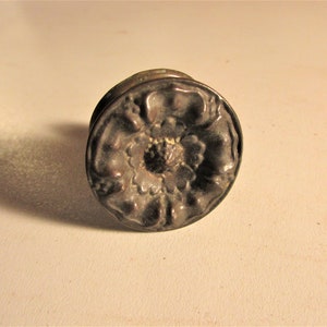 May include: A detailed, antique-style metal knob with a floral design. The knob is circular, with a raised, textured pattern of petals and a central, textured core. The color is a dark, aged bronze.