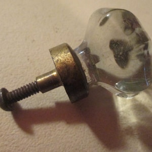 May include: A clear, faceted glass cabinet knob featuring a multi-sided, geometric shape. The knob is attached to a tarnished, weathered brass-toned metal base and a threaded metal screw bolt extending from the back for installation purposes.