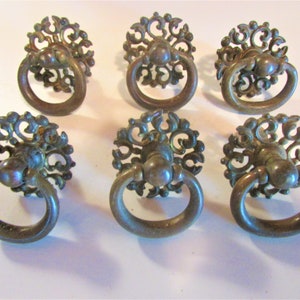 May include: Six antique bronze-colored drawer pulls with ring handles. Each pull features an ornate, floral-like design around the base of the ring. The pulls are arranged in two rows of three, showcasing their vintage aesthetic and potential for furniture restoration.