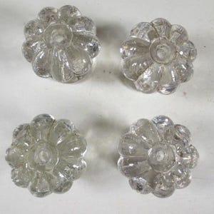 May include: Four clear glass flower-shaped cabinet knobs. Each knob has a central hole and petal-like details. The knobs are arranged in a square pattern against a white background. These are suitable for furniture restoration.