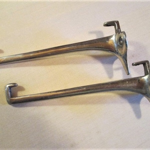 May include: Two vintage, silver-toned metal coat hooks. Each hook has a long, curved arm with a hook at one end and a decorative, bird-like head at the other. The hooks are designed for wall mounting.