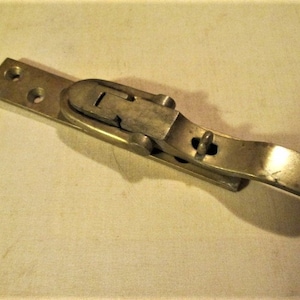 May include: A vintage brass-toned latch with a lever and a rectangular base. The latch has a curved handle and a locking mechanism. The base has two holes for mounting. The metal has a slightly aged appearance.