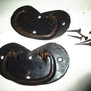 May include: Two antique black metal drawer pulls with a curved handle design. Each pull has a dark, aged appearance with visible rust. Several small, rusty nails are scattered nearby, suggesting a restoration project.
