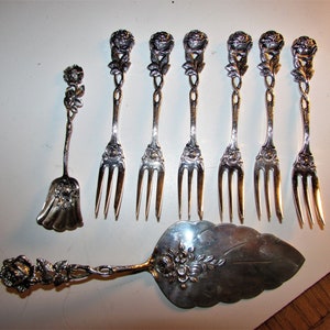 May include: A collection of antique silver serving utensils, including forks and a cake server. The handles are intricately designed with rose motifs. The pieces are arranged on a white surface, showcasing their ornate details.