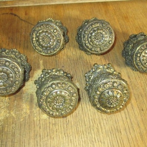 May include: Six antique brass cabinet knobs with a floral design. The knobs have a raised, circular center with a smaller, concentric circle in the center. The knobs are surrounded by a ring of smaller, raised circles.