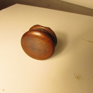 May include: A small, round, dark brown wooden spool with a smooth, polished finish. The object features a cylindrical center section flanked by two circular, flat-faced ends, showcasing a visible wood grain pattern throughout the entire piece.