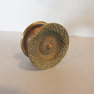 May include: A vintage brass-colored decorative object with a circular, patterned design. The object has a central, raised, textured area surrounded by concentric circles of intricate patterns. The piece appears to be made of metal and has a warm, aged patina.