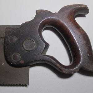 May include: A vintage hand saw with a wooden handle. The handle is dark brown and has a unique shape. The saw blade is made of metal and has a serrated edge.