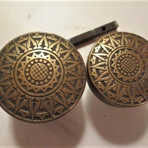May include: Two antique brass cabinet knobs with a sunflower design. The knobs are round with a detailed pattern of petals and a central circular design. A metal bar is visible between the knobs.