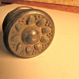 May include: Antique bronze door knob with a floral design. The knob features a central circular element surrounded by stylized petal shapes. The metal has a weathered appearance, suggesting age and use. A classic hardware piece.