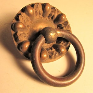 May include: Antique bronze door pull with a circular ring handle. The round backplate features a detailed floral design. The hardware is made of metal and has a vintage aesthetic. The door pull is a decorative element for furniture or doors.