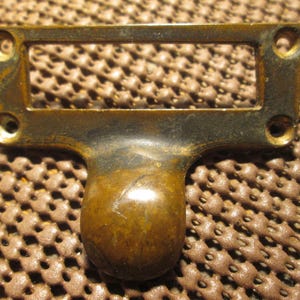 May include: A vintage brass drawer pull with a rectangular label holder and a rounded handle. The brass has a tarnished, aged appearance, with visible wear and discoloration. Four screw holes are present for mounting.