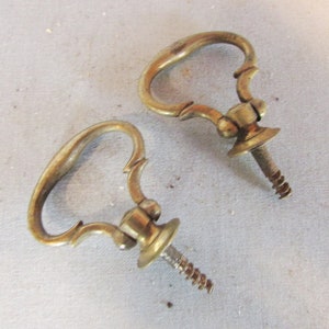 May include: Two antique brass drawer pulls with a curved handle design. Each pull has a decorative base and a screw for attachment. The pulls are a warm gold color and appear to be vintage.