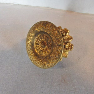 May include: Ornate gold-colored bottle stopper with a floral design. The stopper features a circular top with detailed patterns and a cluster of small flowers and leaves on the side. A decorative item.