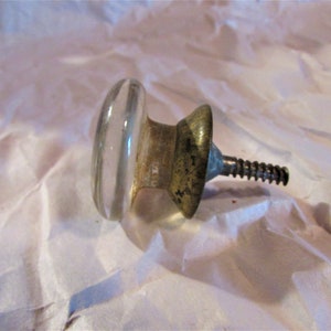 May include: A clear glass and metal cabinet knob with a screw. The knob has a rounded, oval shape and a metal base with a screw for attachment. The metal shows signs of age.