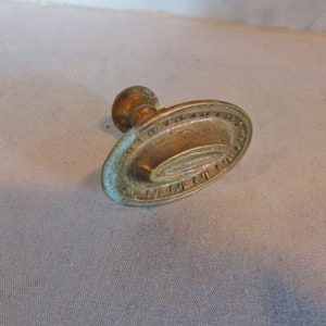 May include: An antique brass drawer pull with an oval backplate and a small, round knob. The backplate has a decorative border and a slightly curved design. The brass has a patina with hints of blue-green oxidation.