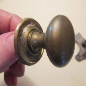 May include: A vintage brass door knob with a rounded, oval-shaped handle and a circular mounting plate. The metal surface shows signs of age, including a darkened patina and some light wear along the edges of the circular base plate.