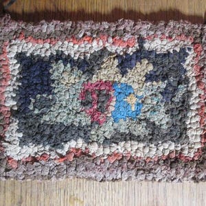 May include: Handmade rag rug with a rectangular shape. The rug features a central design with blue, pink, and beige elements, surrounded by a border of mixed colors. The rug has a rustic, textured appearance, and is likely made from recycled fabric.