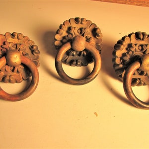 May include: Three antique-style wooden drawer pulls with ring handles. Each pull features a decorative floral design around the base and a circular ring handle. The wood has a warm, aged appearance.