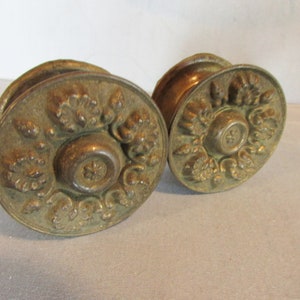 May include: Two antique brass door knobs with floral and scrollwork detailing. The knobs have a round shape with a central rosette design. The brass has a warm, aged patina, suggesting vintage craftsmanship.