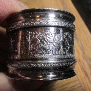 May include: A silver napkin ring with an embossed design. The ring features a detailed scene with figures, trees, and a building. The top and bottom edges have a decorative beaded border. The ring is held by a person.