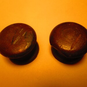 May include: Two dark brown, wooden cabinet knobs with a rounded top and a flat base. The knobs are set against a warm, yellow-orange background, and the wood grain is visible.
