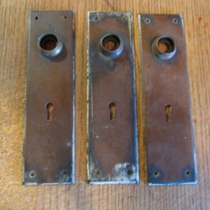 May include: Three antique bronze door plates with keyholes and circular openings for doorknobs. The rectangular plates show signs of age and wear, with a weathered patina and some paint loss, suggesting they are vintage or salvaged.