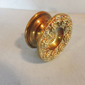 May include: A vintage brass door knob with an ornate floral design. The knob has a circular shape with a detailed pattern around the edge and a central, smaller circular design. The brass has a warm, golden hue.