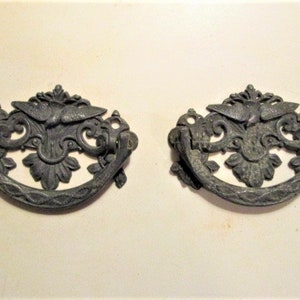 May include: Two antique drawer pulls with a dark gray finish. Each pull features an ornate design with a central bird motif, floral accents, and a curved handle. These decorative pulls are likely for furniture restoration.