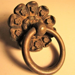 May include: A bronze-colored door pull with a floral design. The pull features a large, flower-shaped base with smaller flower details and a circular handle. The hardware is likely made of metal and is designed for a door or drawer.