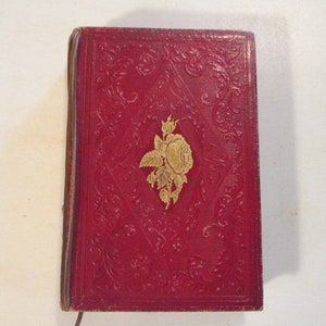 May include: A red leather-bound book with embossed floral patterns and a gold-colored rose emblem. The book's cover is a deep red, and the edges show signs of wear, suggesting age.