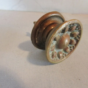 May include: Antique bronze-toned cabinet knob with an oval face featuring a floral design. The knob has a detailed, aged appearance, with a central rounded element and surrounding petal-like shapes. The hardware is likely for furniture or cabinetry.