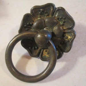 May include: Antique bronze-toned drawer pull with a floral backplate and a circular handle. The flower-shaped backplate has detailed petal designs. The handle is a simple, rounded ring. This hardware is suitable for cabinets and drawers.