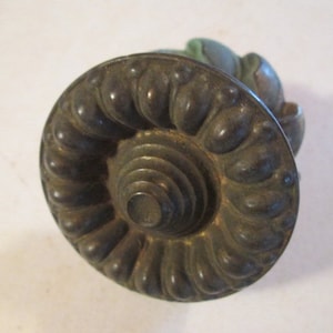 May include: Antique bronze-toned cabinet knob with a spiral and floral design. The knob has a circular shape with a detailed, textured surface. The design features a central spiral surrounded by a series of rounded, petal-like shapes.