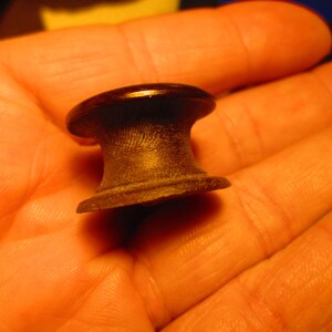 May include: A small, dark brown wooden knob with a rounded top and a flared base. The knob is held in a hand, and the wood grain is visible. The knob appears to be a furniture component.
