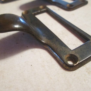 May include: A vintage brass drawer pull with a long, curved handle and a rectangular plate with a slot for a label. The pull has a dark patina finish.