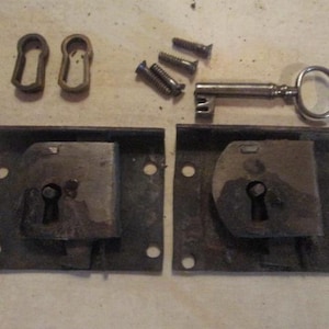 May include: An assortment of antique hardware components, including two metal lock boxes, a key, screws, and keyhole escutcheons. The metal pieces have a weathered, aged appearance, suggesting they are vintage or salvaged items.