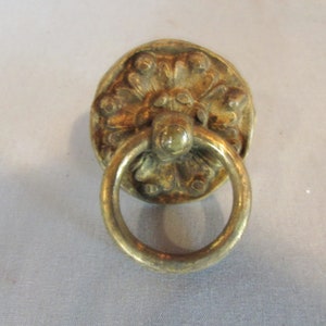 May include: Antique brass drawer pull with a circular, floral-patterned backplate and a rounded ring handle. The hardware has a weathered, aged appearance, suggesting vintage or antique origins. The pull is designed for furniture or cabinetry.