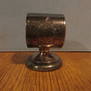 May include: A silver-colored napkin ring with a floral design. The ring is cylindrical with a decorative pattern and sits on a small, round base. The napkin ring is made of metal and is displayed on a wooden surface.